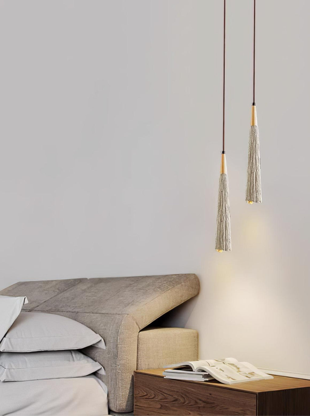 Two narrow Clérachic pendants glow just above a taupe headboard and a wooden nightstand with an open magazine.