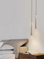 Two narrow Clérachic pendants glow just above a taupe headboard and a wooden nightstand with an open magazine.