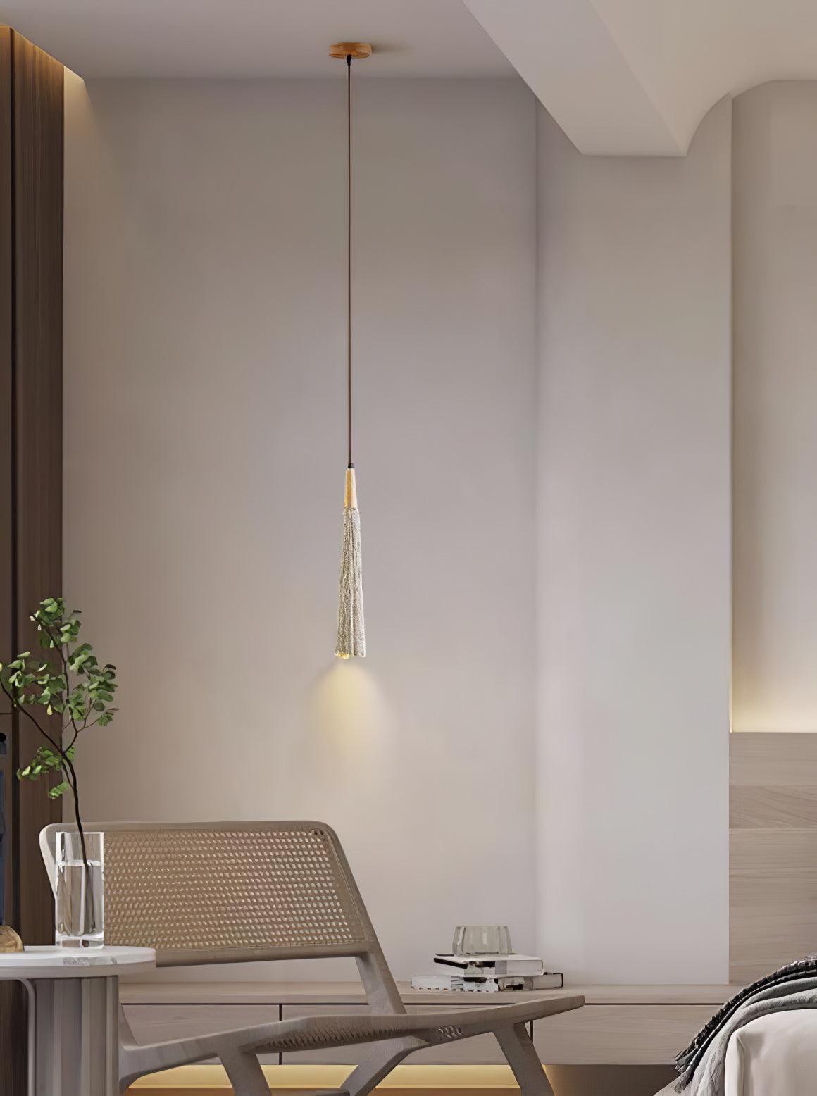 One slim Clérachic pendant hangs by a rattan lounge chair, with a small plant on a side table in the corner.
