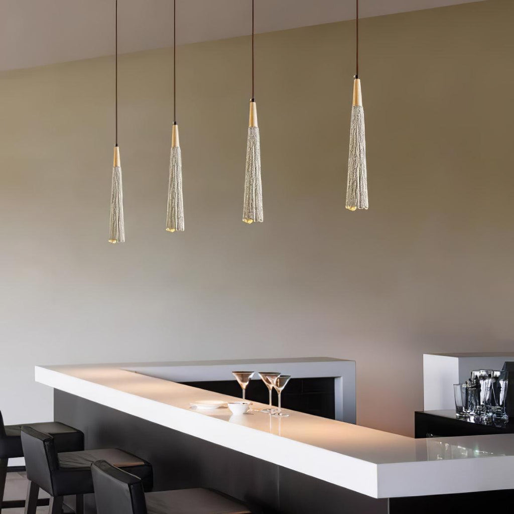Four slim Clérachic pendants hang in a line over a white bar counter set with cocktail glasses and black stools.
