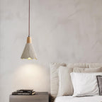 A bell-shaped Clérachic pendant hangs over a dark nightstand with a stack of books, beside a bed with white pillows.