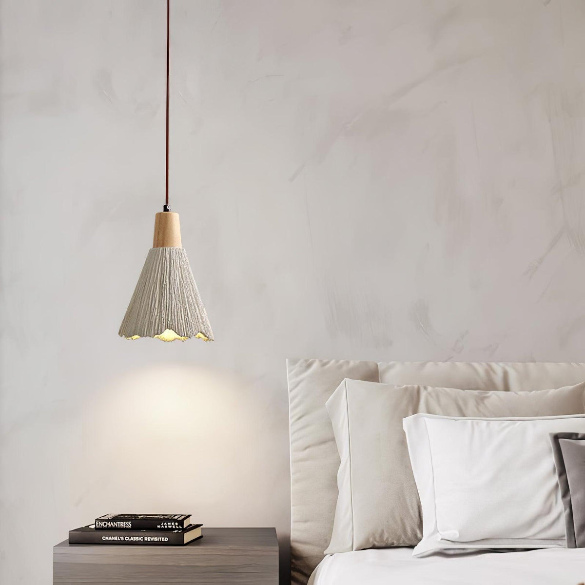 A bell-shaped Clérachic pendant hangs over a dark nightstand with a stack of books, beside a bed with white pillows.
