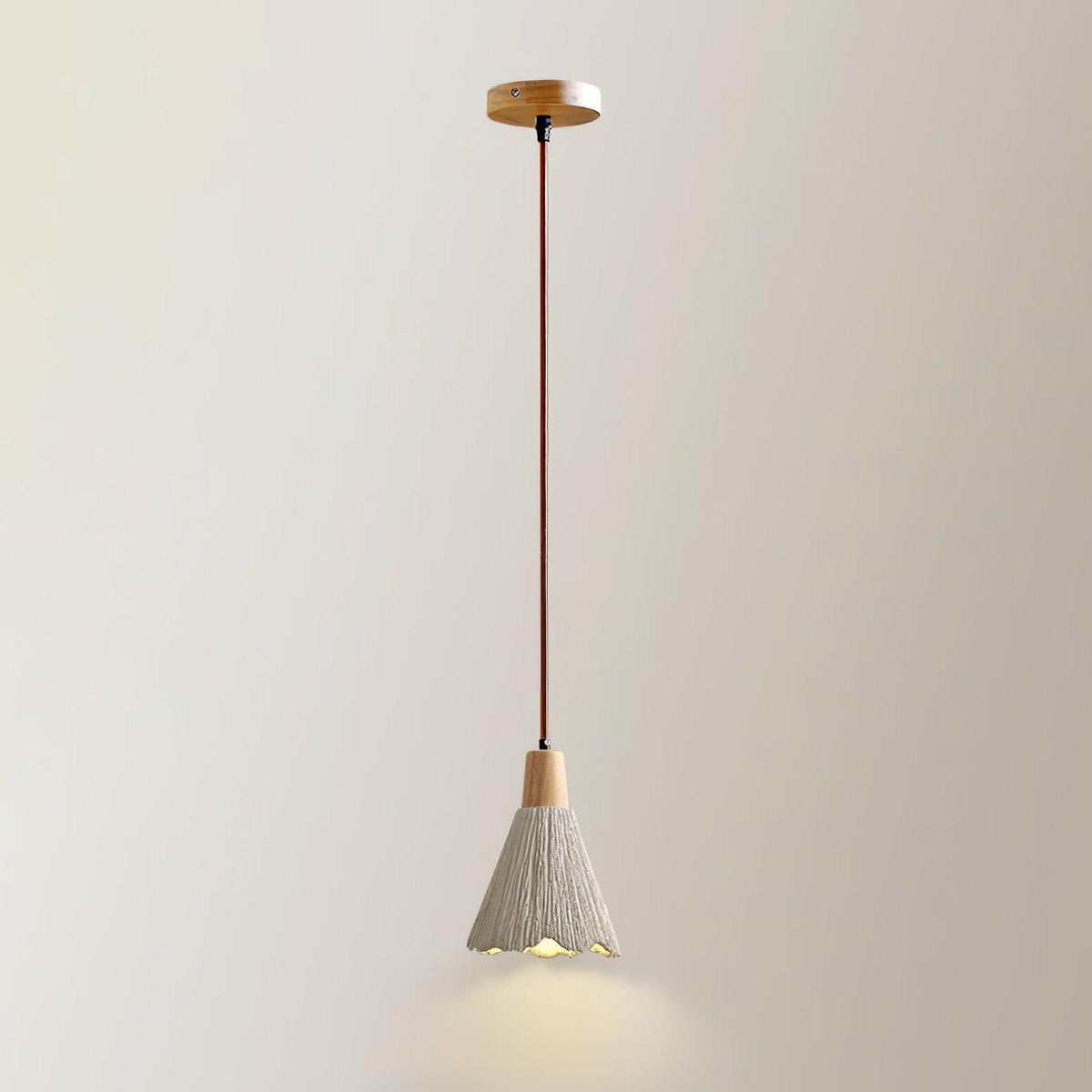 A Clérachic pendant with a bell shade hangs from a round wooden canopy against a plain cream wall, its rim lit warm.
