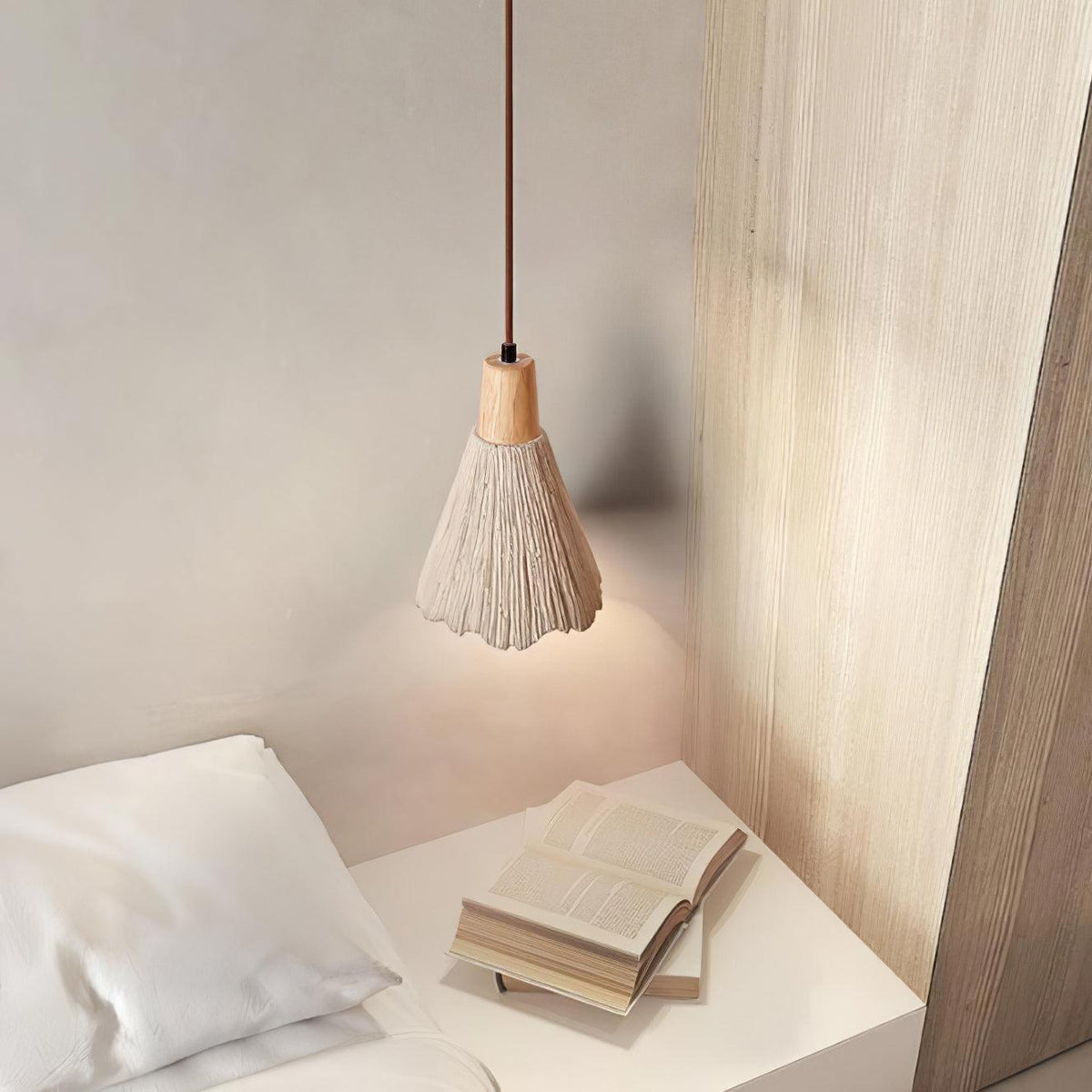A small bell-shaped Clérachic pendant hangs over a white bedside table with an open book, beside a pale wood panel.
