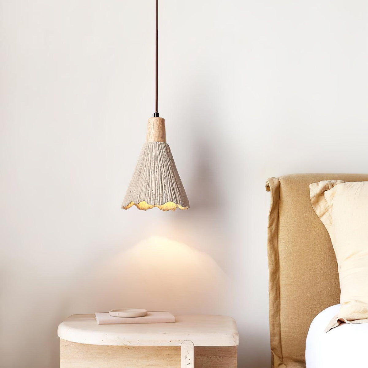 A Clérachic pendant with a bell shade hangs over a round stone side table, a golden bedhead behind it.