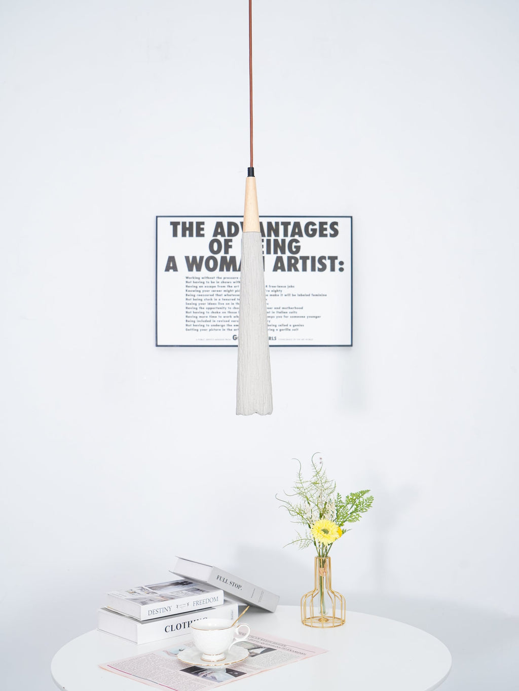 A slim Clérachic pendant hangs in front of a framed print above a small round table with books and a vase of flowers.