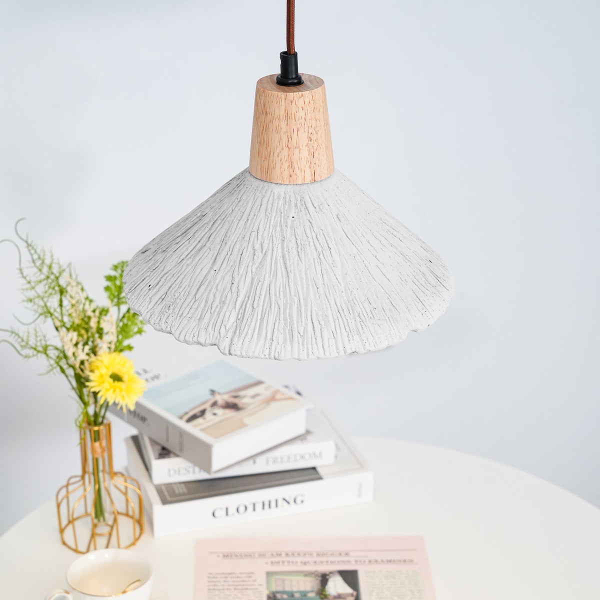 Only the underside of this Clérachic pendant shows over a white table with stacked books and a vase of yellow flowers.