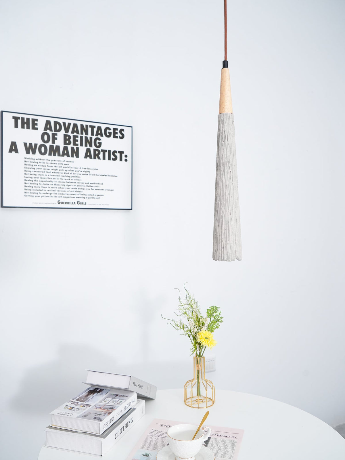 A narrow Clérachic pendant hangs beside a framed print, over a white table with books, a teacup and a gold vase of flowers.