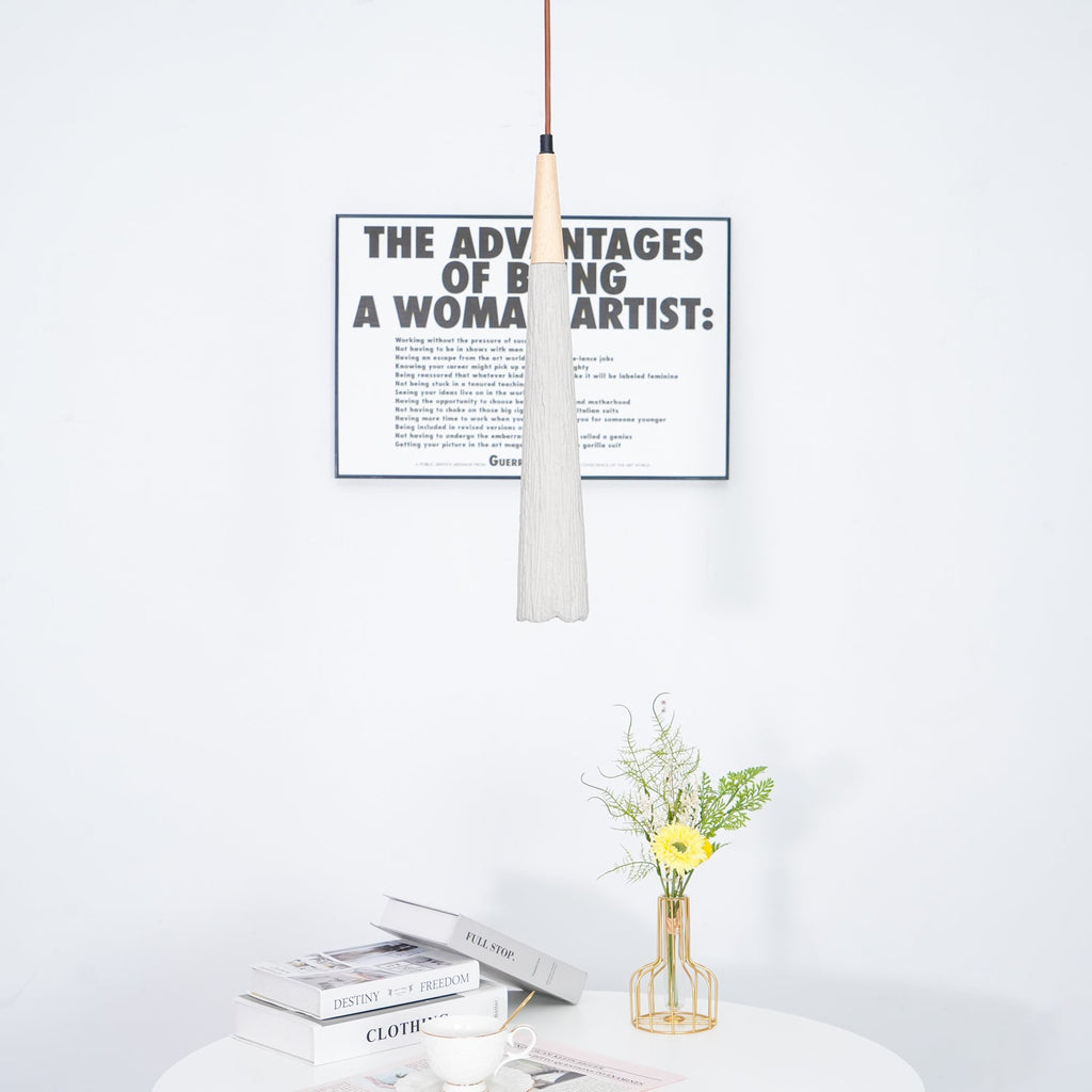 A slim grey Clérachic pendant with a wooden collar hangs in front of a framed print, over a table with books and flowers.