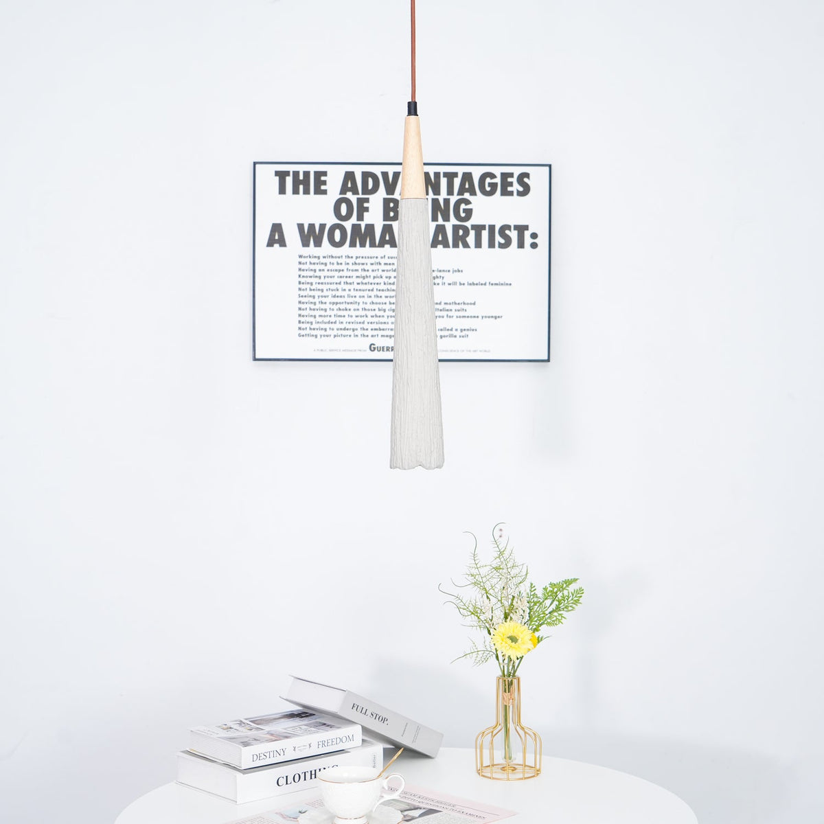 A slim grey Clérachic pendant with a wooden collar hangs in front of a framed print, over a table with books and flowers.