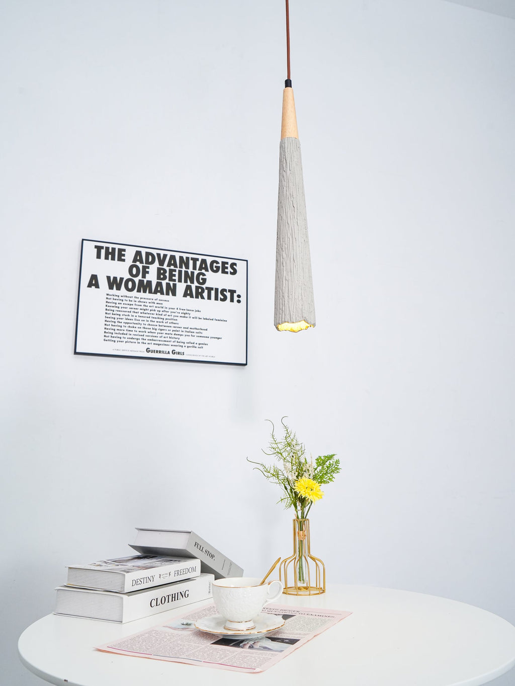 A slim Clérachic pendant glows at the tip, with a framed print behind it and books, a cup and flowers on a table below.