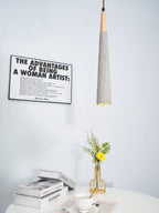 A narrow Clérachic pendant hangs in front of a framed print, above books, a teacup and a gold vase of yellow flowers.