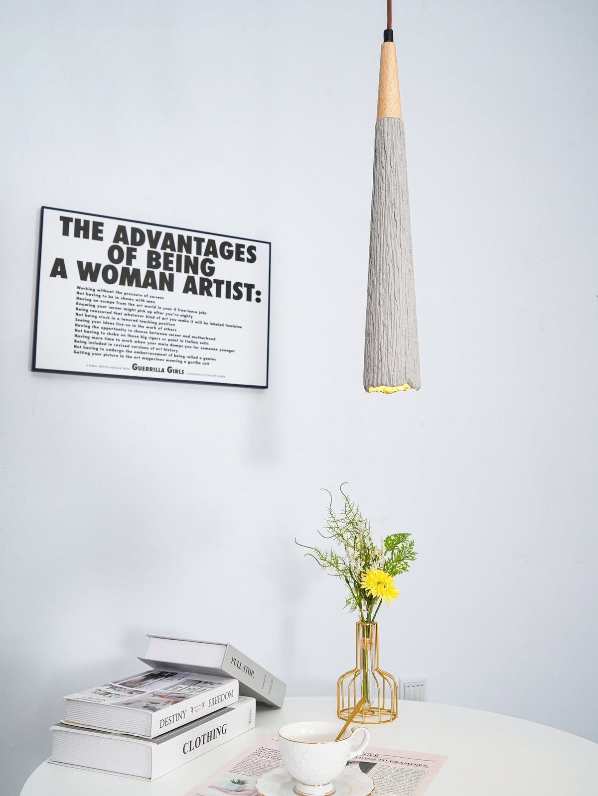 A narrow Clérachic pendant hangs in front of a framed print, above books, a teacup and a gold vase of yellow flowers.