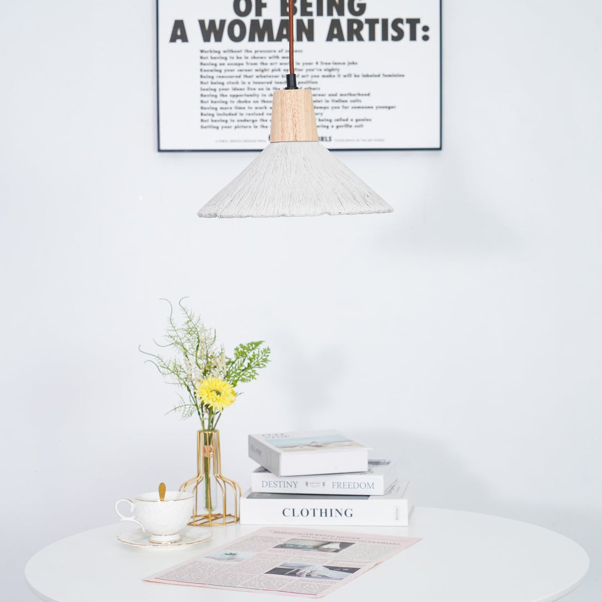 Part of a wide-shade Clérachic pendant at the top of the frame, with a framed print behind and a table of books and flowers below.
