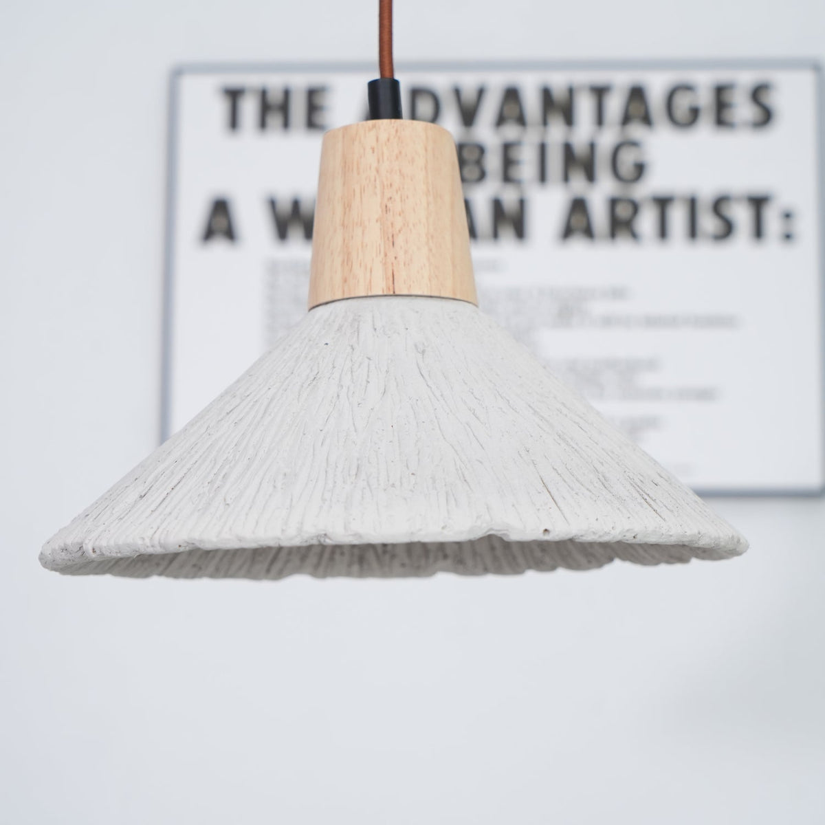 Wide grey Clérachic pendant in front of a blurred framed print, its wooden cap and ridged shade in sharp focus.