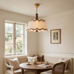 A Clérachic pleated shade with wavy brown trim lit above a round wooden table in a breakfast nook beside a sash window.