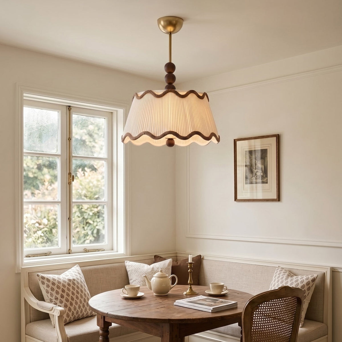 A Clérachic pleated shade with wavy brown trim lit above a round wooden table in a breakfast nook beside a sash window.