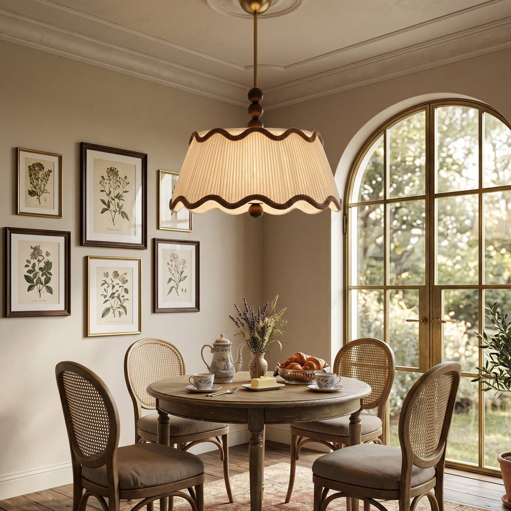 Lit pleated Clérachic ceiling light above a round dining table with cane-backed chairs, near an arched window and framed botanical prints.