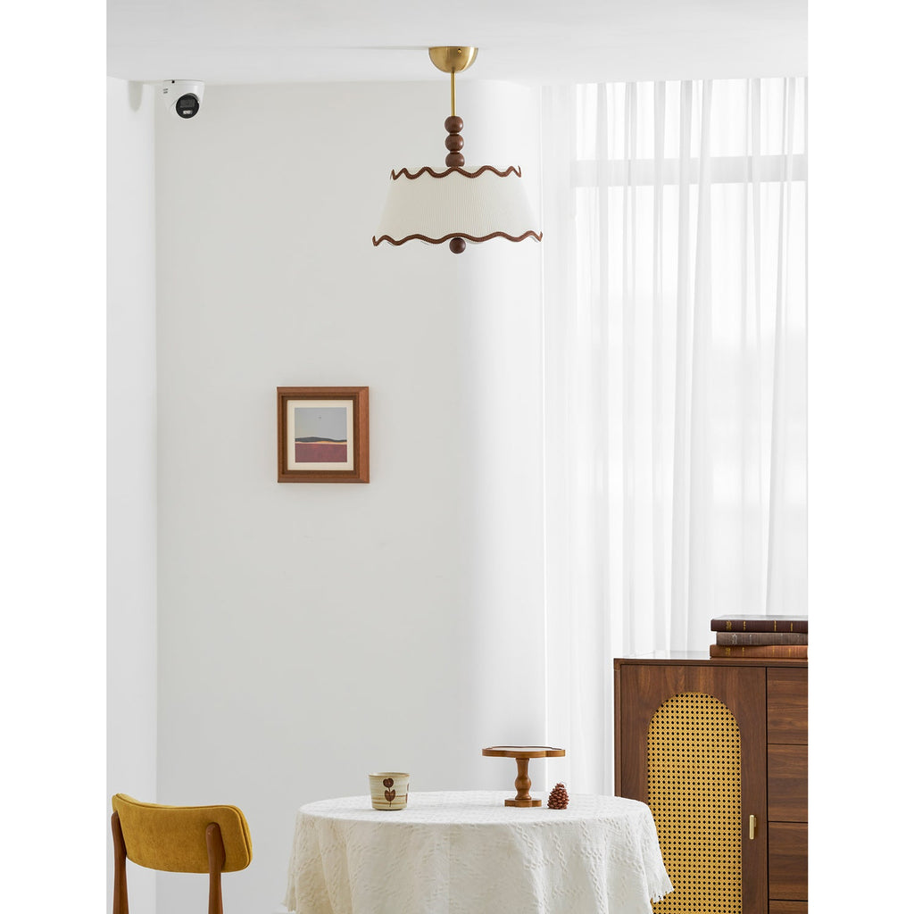 A Clérachic pleated ceiling light seen from a distance in a white room, with sheer curtains, a cane-fronted cabinet and a small round table.
