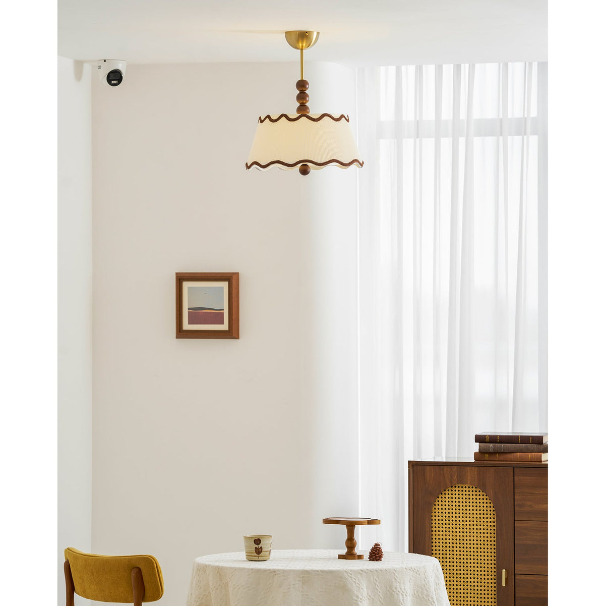 Wider view of the Clérachic pleated ceiling light in a white room with sheer curtains, a cane-fronted wooden cabinet and a round table.
