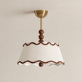 Close view of the wavy brown trim, pleated white shade, wood beads and brass canopy of this Clérachic ceiling light against a beige wall.