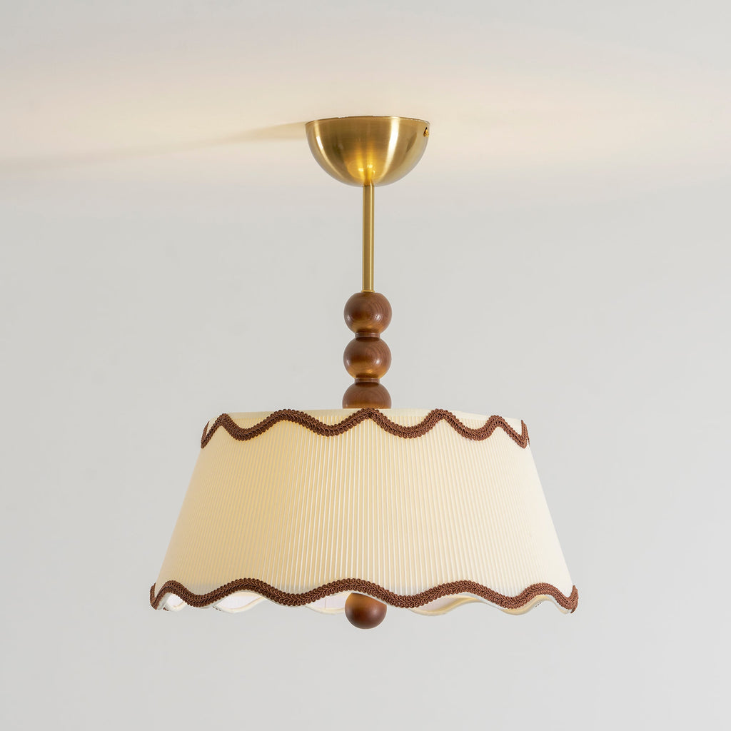 Clérachic pleated ceiling light in a warm cream shade with wavy brown braid trim, stacked wood beads and a brass canopy, against a pale wall.