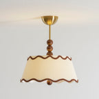 Clérachic pleated ceiling light in a warm cream shade with wavy brown braid trim, stacked wood beads and a brass canopy, against a pale wall.