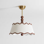 Clérachic ceiling light with a white pleated tapered shade, wavy brown trim and a brass canopy, against a light grey ceiling and wall.