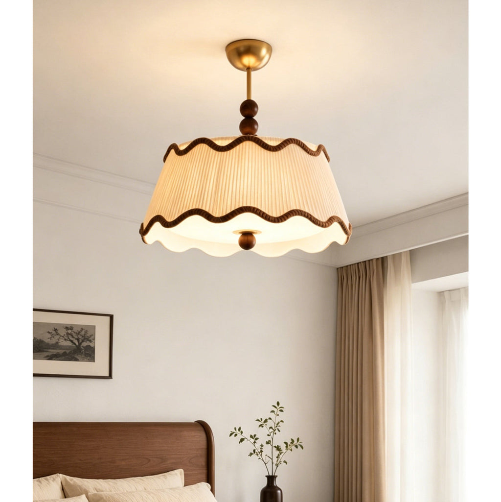 A Clérachic pleated ceiling light glowing warm above a bed with a walnut headboard, next to a framed landscape print and pale curtains.