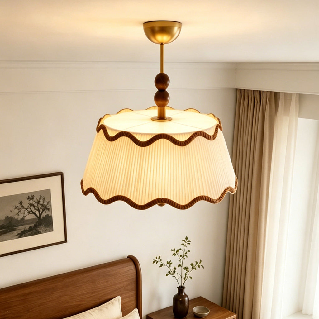 View from below of the lit Clérachic pleated shade and walnut beads, with a wooden bed and a vase of leafy stems nearby.