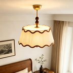 View from below of the lit Clérachic pleated shade and walnut beads, with a wooden bed and a vase of leafy stems nearby.