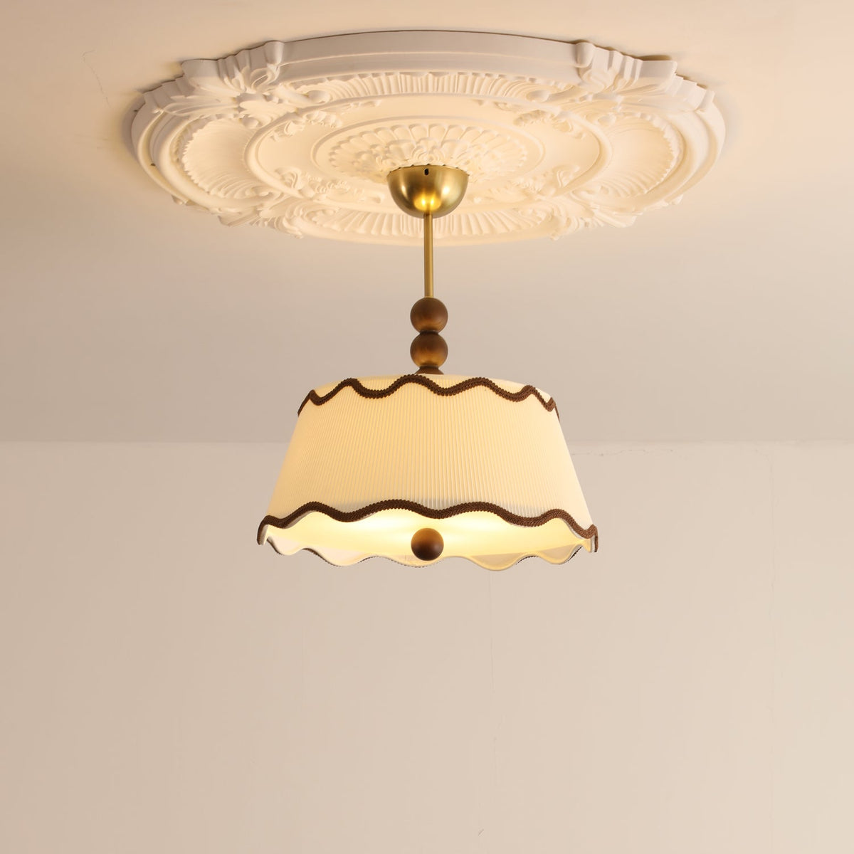 The same Clérachic pleated ceiling light lit warm, hanging beneath a decorative plaster ceiling rose.