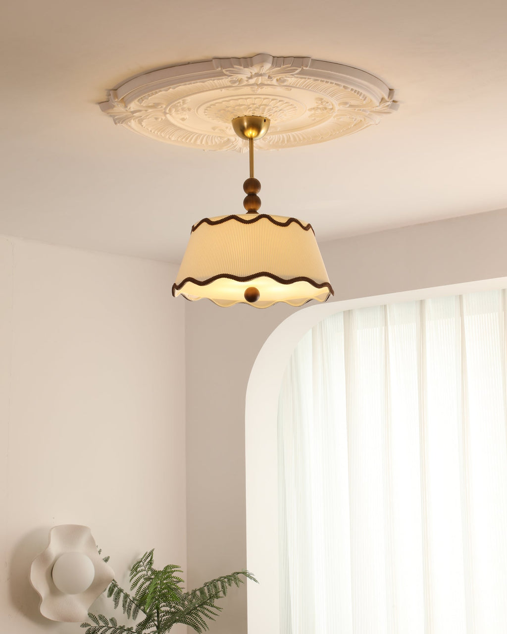Lit Clérachic ceiling light under a plaster rose, with an arched white wall, sheer curtains and a green fern in the corner.