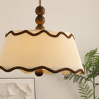 Close view of the pleated cream shade and scalloped brown trim of this Clérachic ceiling light, with a fern and a framed print behind.