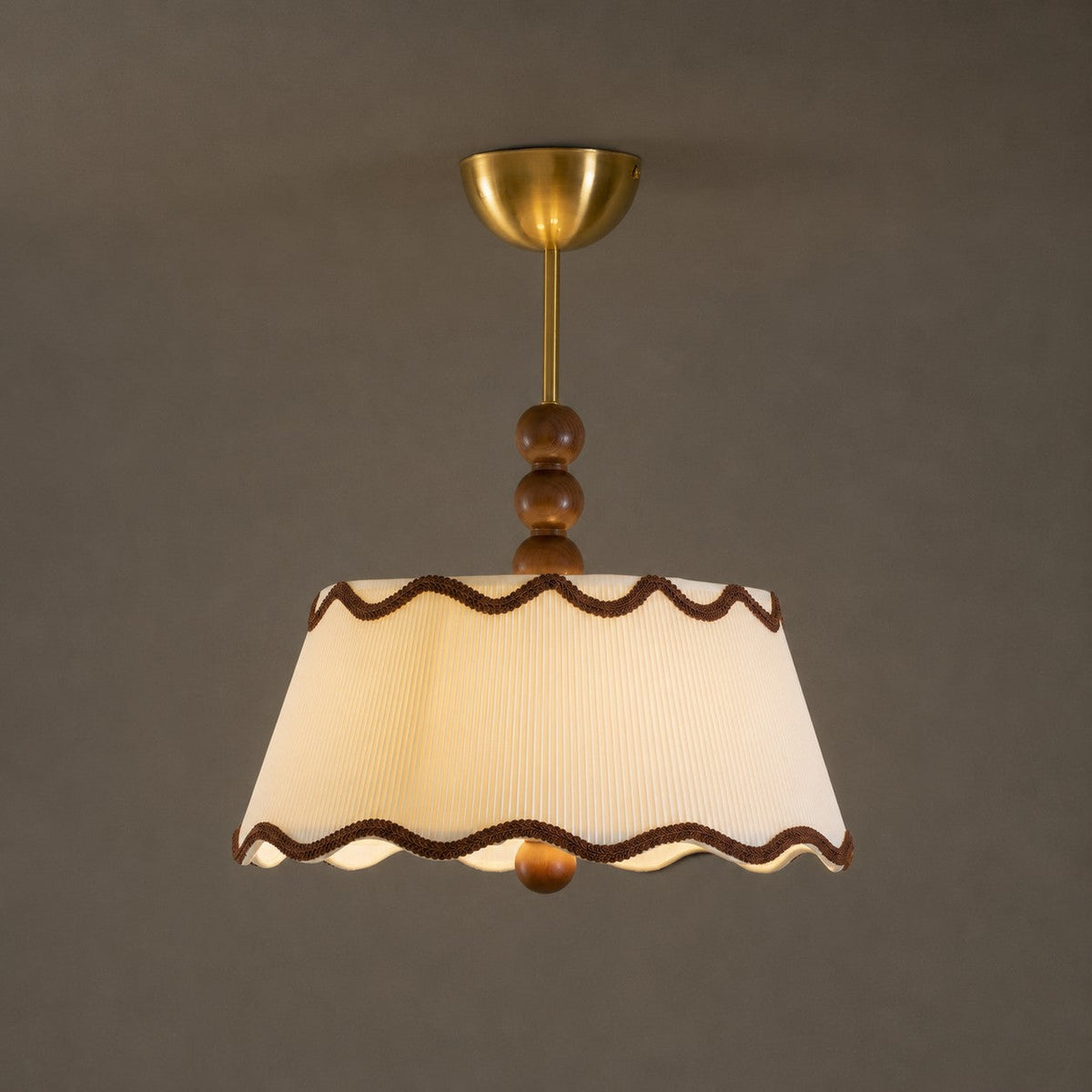 Lit Clérachic pleated ceiling light with brown trim and a brass canopy, glowing warm against a dark taupe wall.
