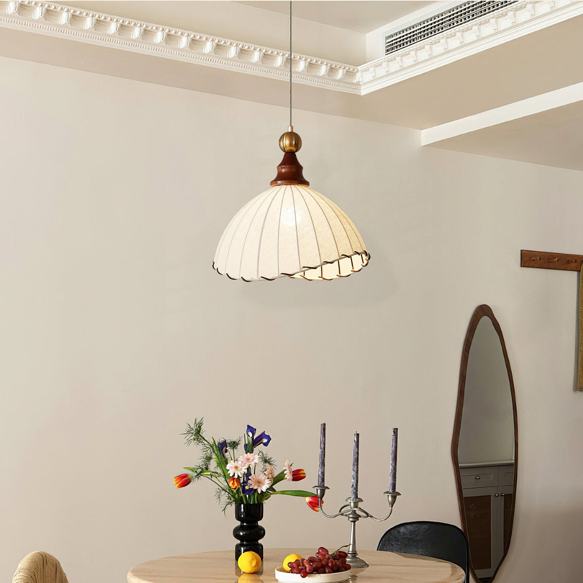 A Clérachic pendant light with a scalloped beige shade, hanging below a ceiling cornice beside an oval mirror and candlesticks.
