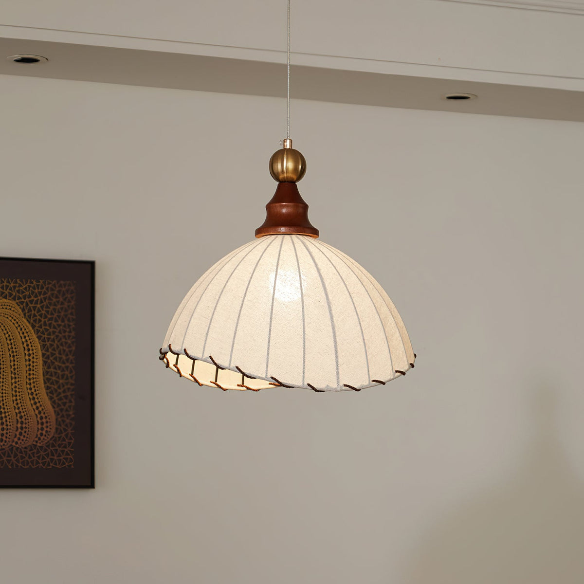 A Clérachic pendant light lit warmly in a grey-walled room, with a framed dark print to its left and a recessed ceiling behind.