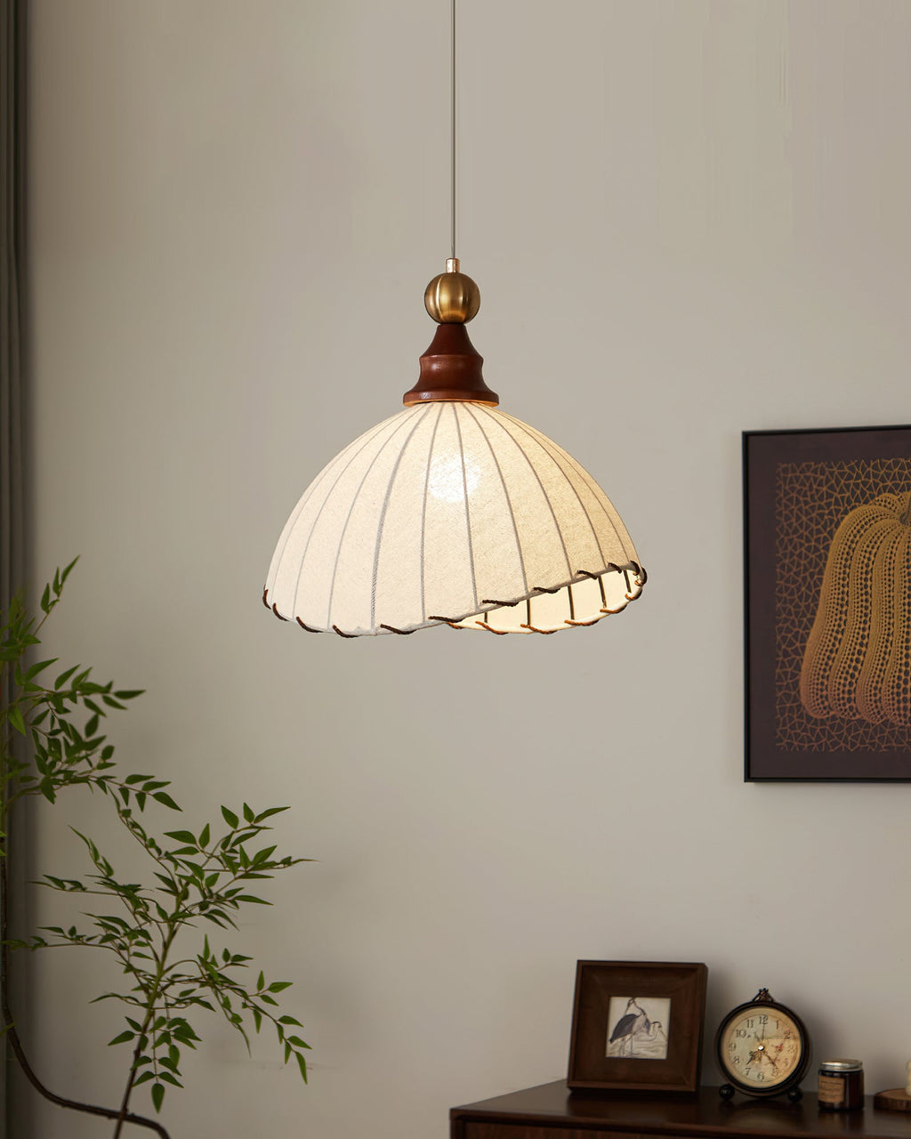A Clérachic pendant light beside a leafy branch and a framed print, with a small clock and photo frame on a cabinet below.