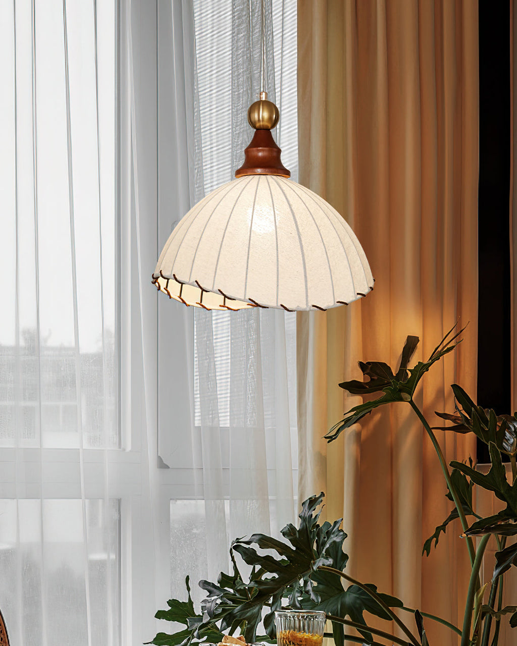 This Clérachic pendant light lit up beside sheer curtains and a gold drape, with large green leaves in the foreground.