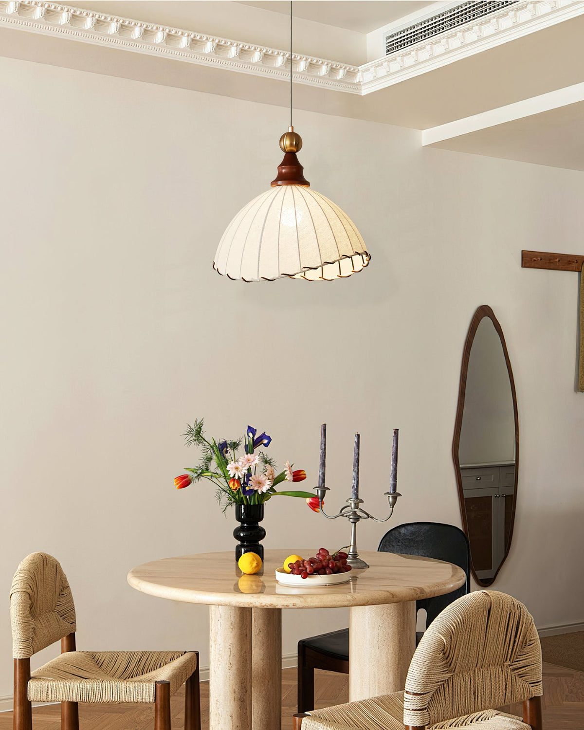 Beige Clérachic pendant light over a round stone table with cane chairs, flowers and candlesticks in a cornice-trimmed room.