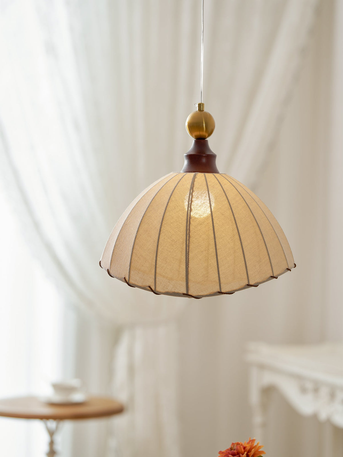Clérachic pendant light seen against sheer white curtains, with its warm light showing through the beige pleated fabric.
