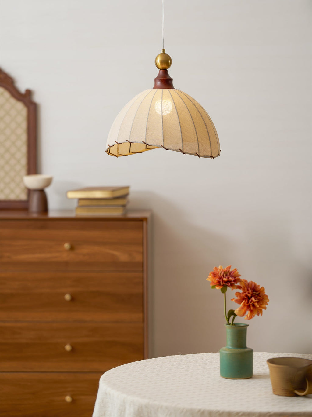 This Clérachic pendant light over a round table with an orange flower in a green vase, beside a walnut chest of drawers.