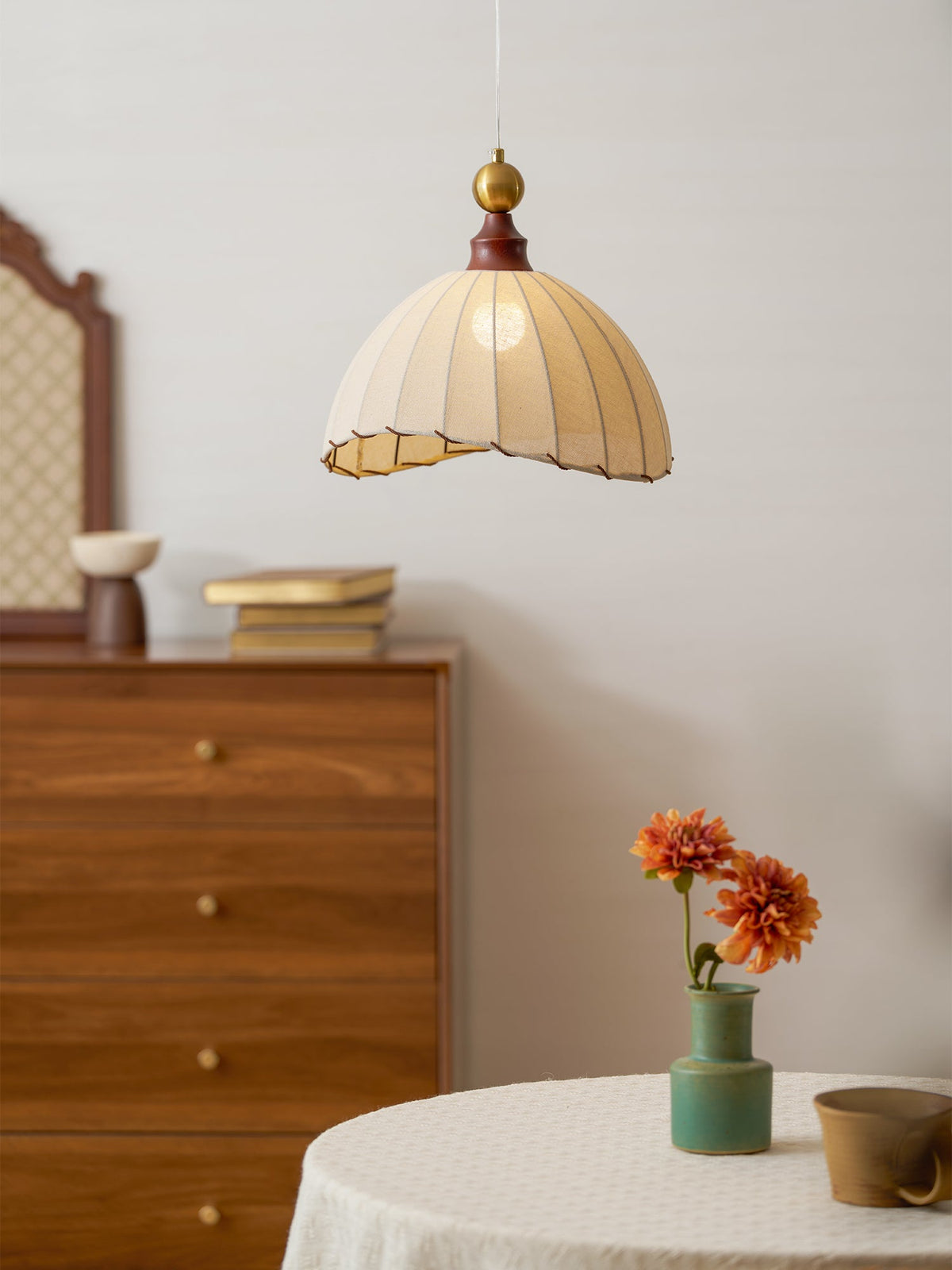 This Clérachic pendant light over a round table with an orange flower in a green vase, beside a walnut chest of drawers.
