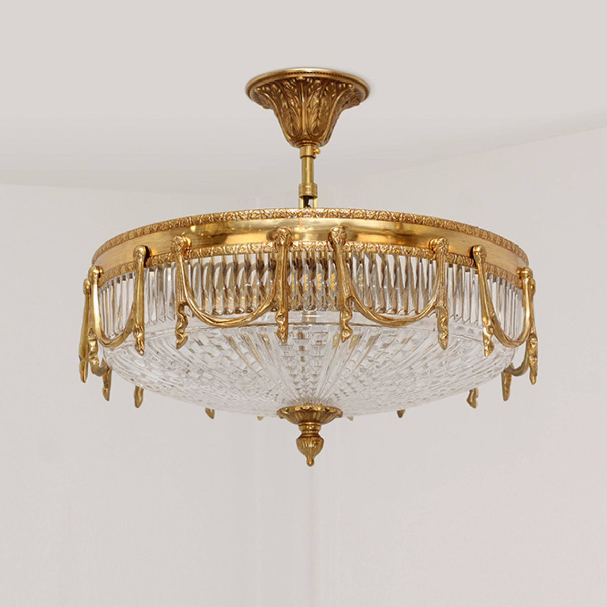 Side-on view of this Clérachic brass ceiling light, with draped brass swags over a pleated clear glass bowl.