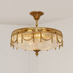 Angled view of the Clérachic brass ceiling light lit, showing the carved brass canopy and the glass ribbing in warm light.