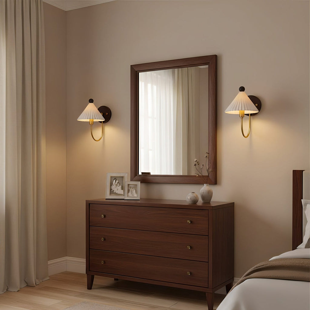 A pair of Clérachic wall lamps with pleated white shades lit either side of a framed mirror above a dark wood chest of drawers.