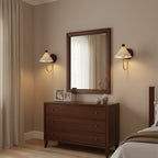 A pair of Clérachic wall lamps with pleated white shades lit either side of a framed mirror above a dark wood chest of drawers.