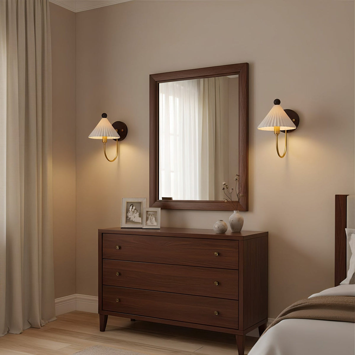 A pair of Clérachic wall lamps with pleated white shades lit either side of a framed mirror above a dark wood chest of drawers.