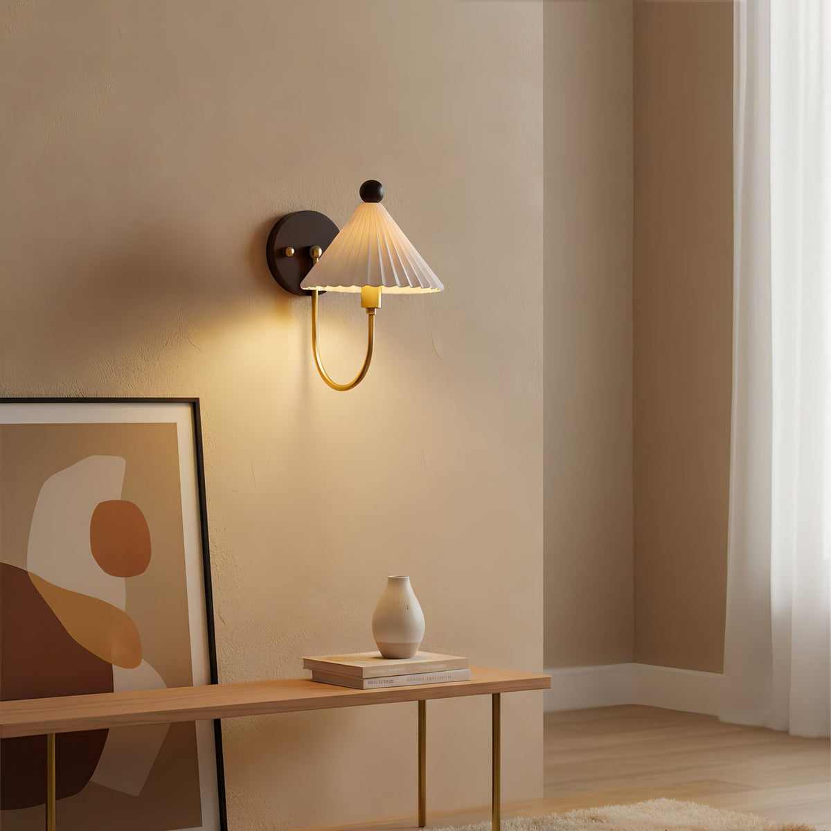 A lit Clérachic wall lamp with a pleated white shade and curved brass arm, above a bench with a vase and a large abstract print.