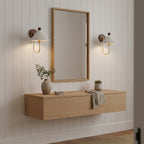 Two Clérachic wall lamps with pleated shades flanking a tall mirror above a floating oak shelf, against white panelled walls.